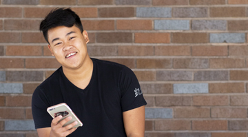 young man in front of brick wall using phone, smiling at camera