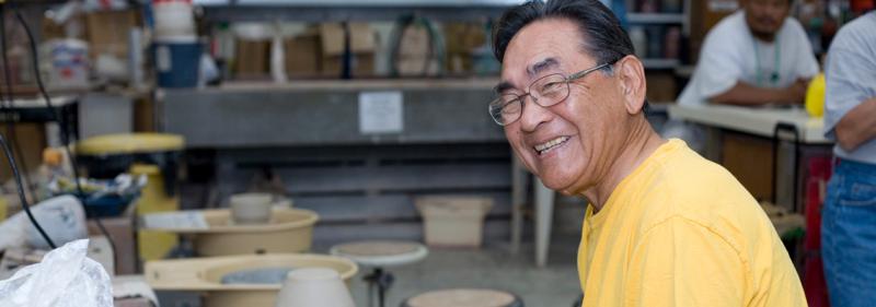 older man sitting at ceramic wheel smiling at camera